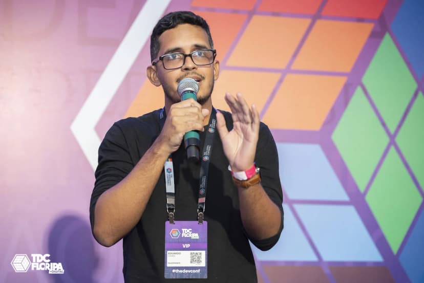 Eduardo Corrêa falando em palco durante evento de tecnologia, com fundo colorido e banner do TDC Floripa, usando blusa preta e óculos, segurando microfone. Eduardo Corrêa falando em palco durante evento de tecnologia, com fundo colorido e banner do TDC Floripa, usando blusa preta e óculos, segurando microfone.