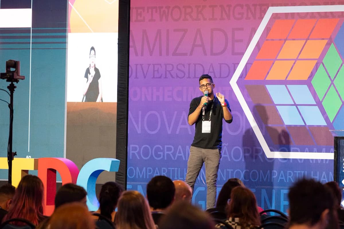 Eduardo palestrando sobre acessibilidade no palco com destaque para as letras TDC em cores vibrantes, em um ambiente com fundo em tons de rosa e roxo, promovendo networking, inovação e programação. Eduardo palestrando sobre acessibilidade no palco com destaque para as letras TDC em cores vibrantes, em um ambiente com fundo em tons de rosa e roxo, promovendo networking, inovação e programação.